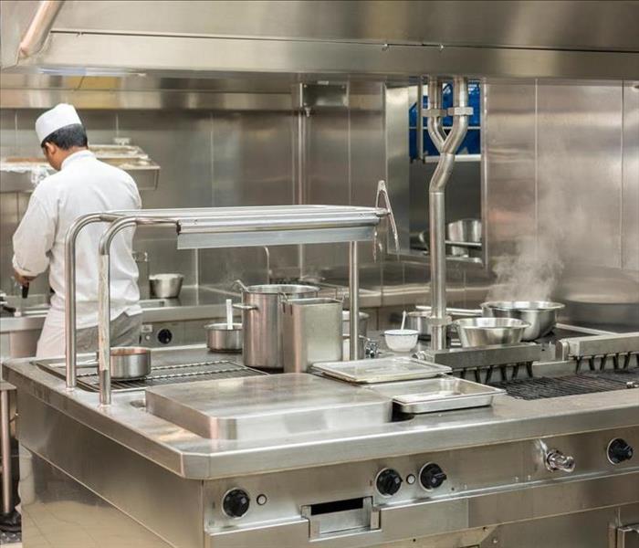 Stainless steel commercial kitchen, with a chef in a white jacket and chefs hat in the background 